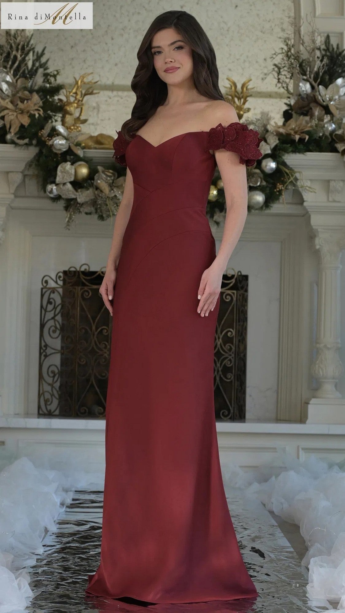 A woman in a wine-colored Rina Di Montella RD3325 long formal dress with an off-the-shoulder sheath silhouette stands indoors before a decorated fireplace.